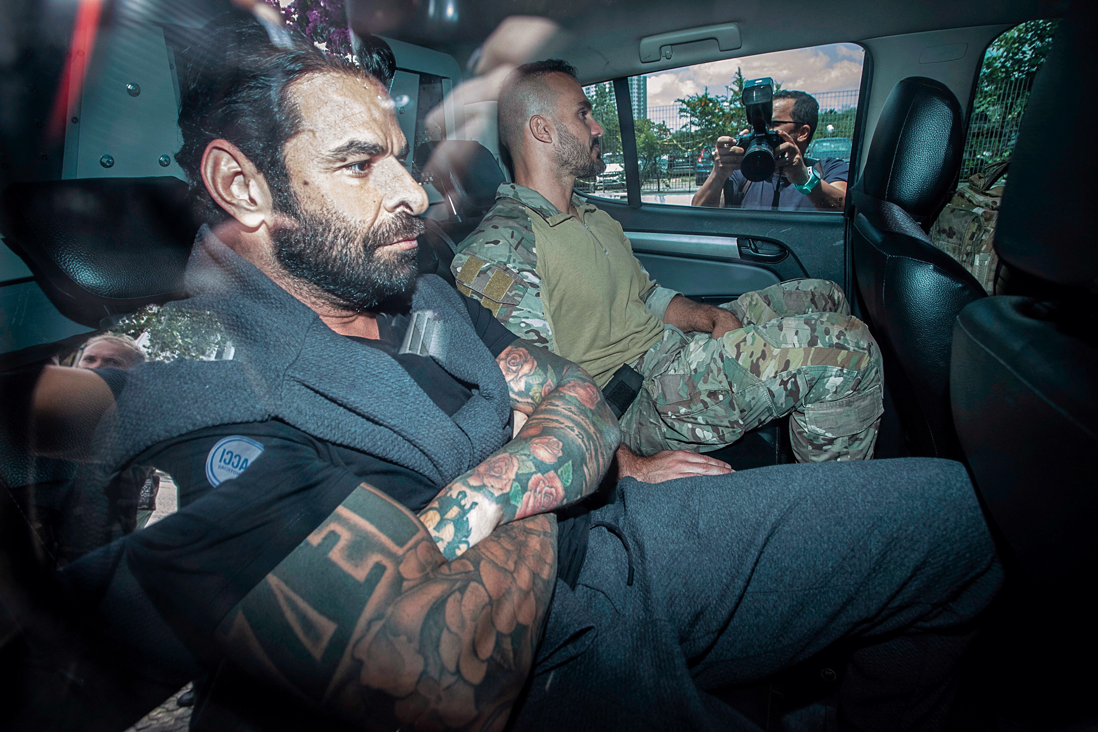 Homem barbudo com braços tatuados e blusa cinza sentado no banco de trás de um carro, ao lado de outro homem de uniforme camuflado. Um fotógrafo com câmera profissional aparece na janela do veículo
