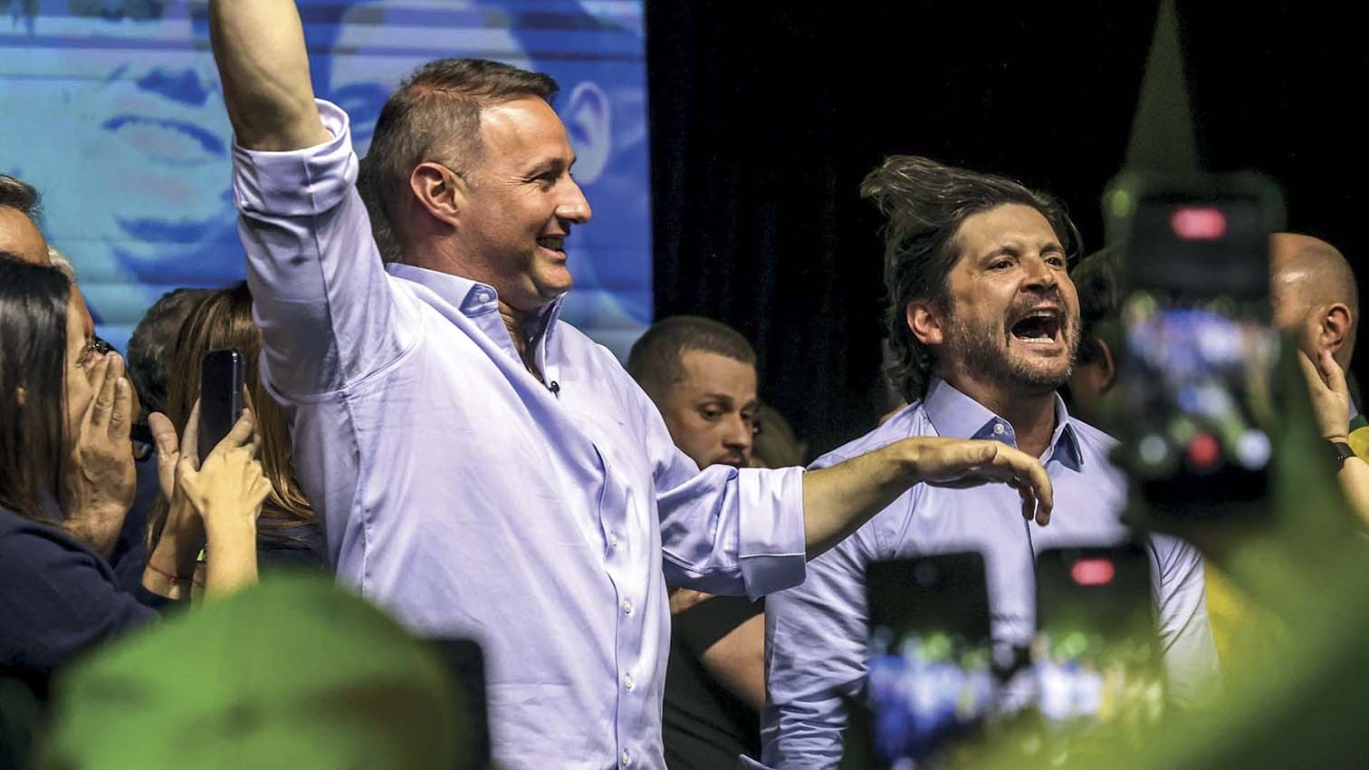 Dois homens em camisas sociais claras, um com o braço erguido e sorrindo, outro gritando com a boca aberta, em meio a uma multidão com celulares filmando