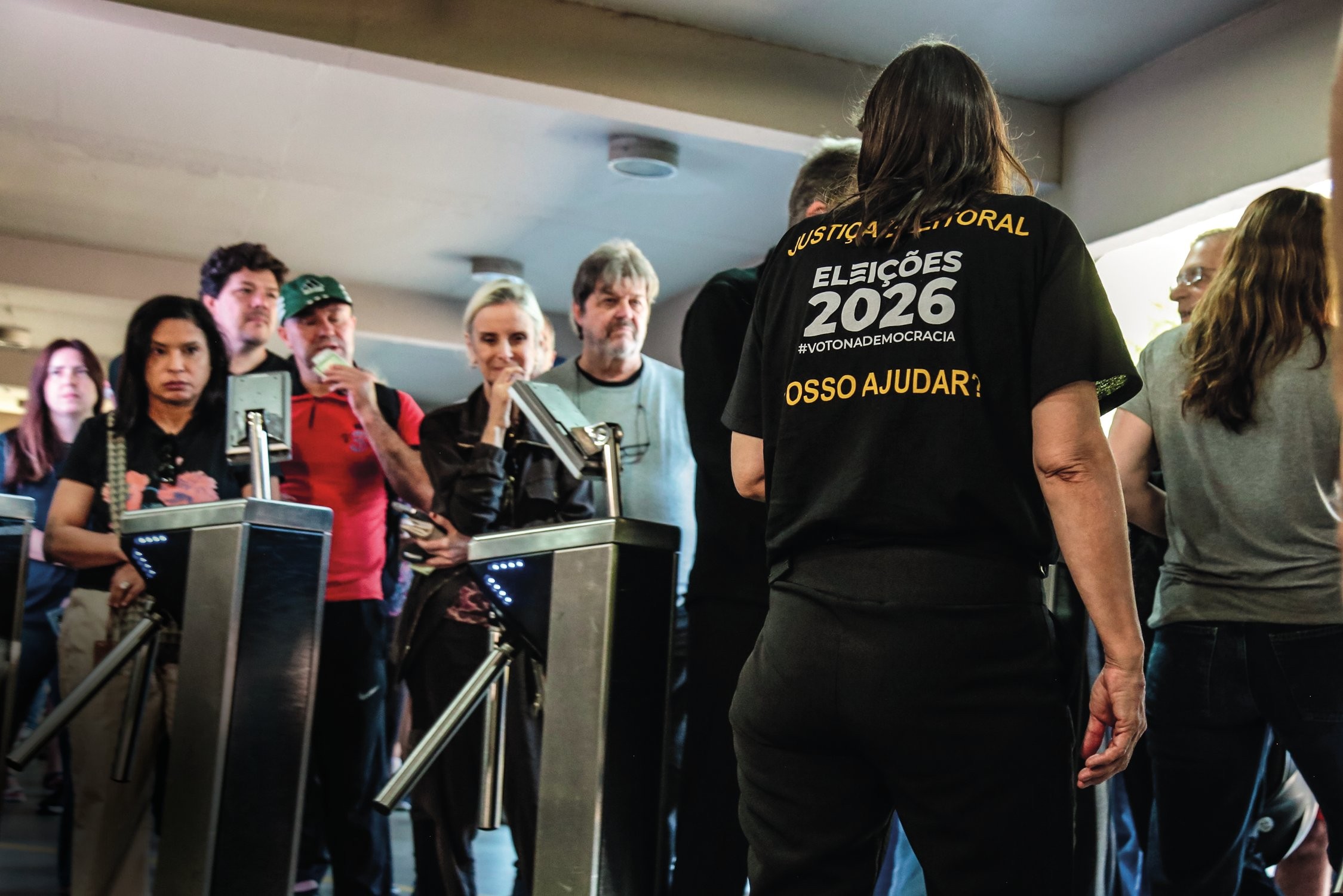 Pessoas em uma fila, com catracas eletrônicas à frente. Uma mulher de costas veste camiseta preta com JUSTIÇA ELEITORAL ELEIÇÕES 2026 #VOTONADEMOCRACIA POSSO AJUDAR? em amarelo. Outros indivíduos aguardam, alguns olhando para a frente, outros conversando