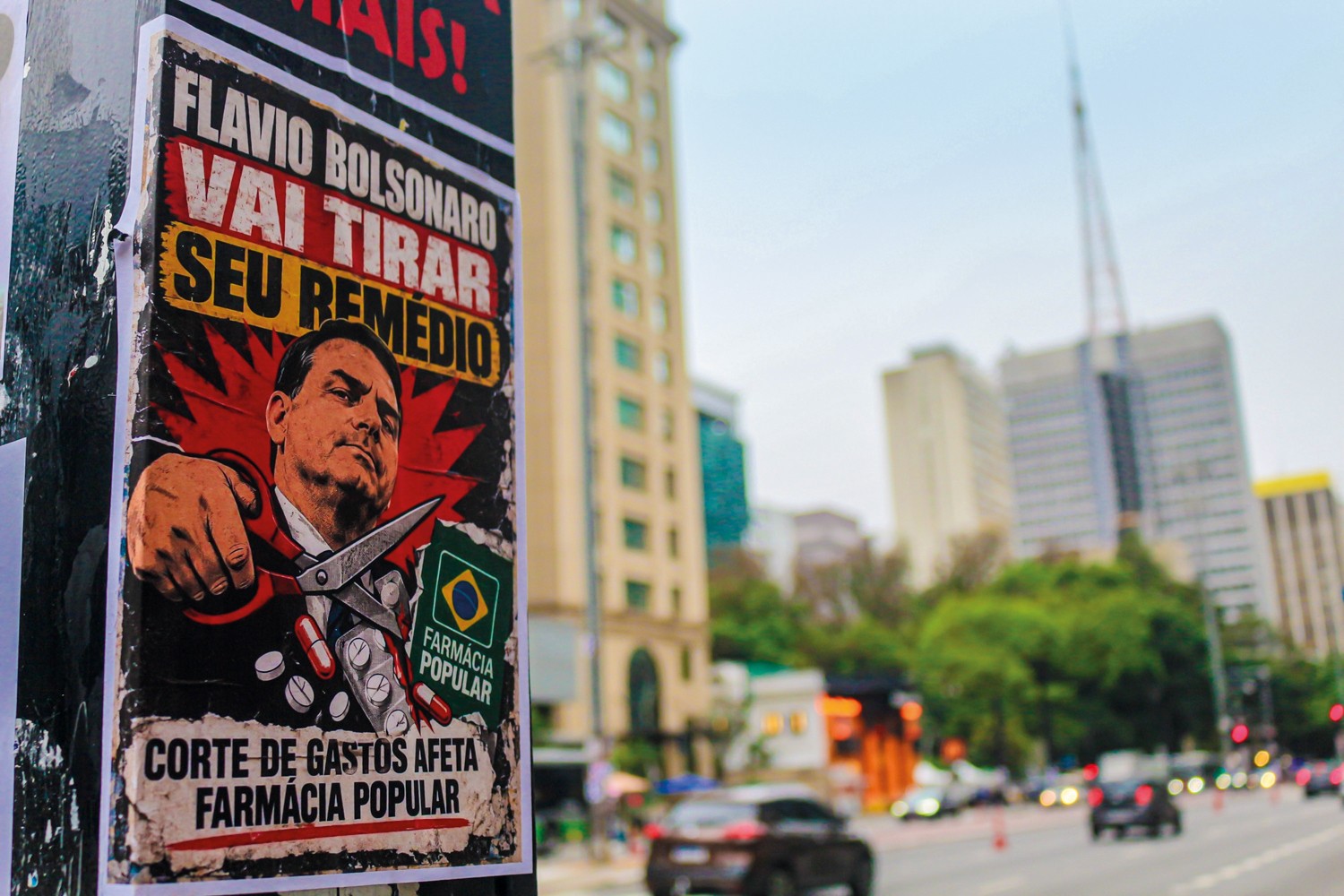 Cartaz político colado em poste, com o rosto de Jair Bolsonaro segurando uma tesoura que corta pílulas e cartelas de remédio. O texto diz FLAVIO BOLSONARO VAI TIRAR SEU REMEDIO e CORTE DE GASTOS AFETA FARMACIA POPULAR. Ao fundo, prédios e uma rua movimentada em dia nublado