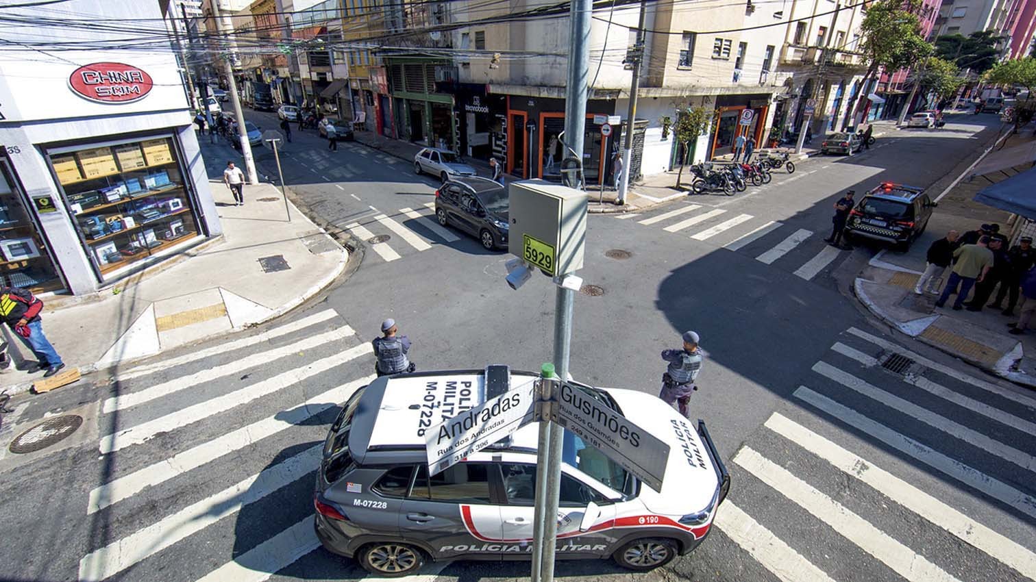 Vista aérea de um cruzamento urbano movimentado com uma viatura da Polícia Militar estacionada e dois policiais próximos. Pessoas caminham nas calçadas e atravessam a rua, com carros e motos estacionados. Edifícios comerciais e residenciais ladeiam as ruas, sob um céu claro