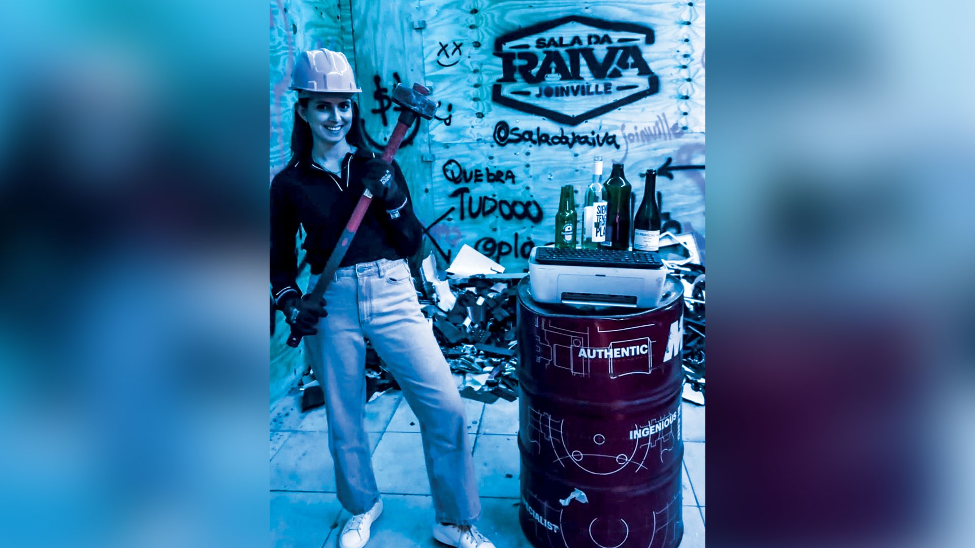 Mulher sorridente, usando capacete de segurança, blusa escura e calça jeans clara, segura uma marreta. Ela está em um ambiente azulado com pichações SALA DA RAIVA JOINVILLE e QUEBRA TUDOOO. Ao lado, barris vermelhos empilhados com garrafas e um teclado.