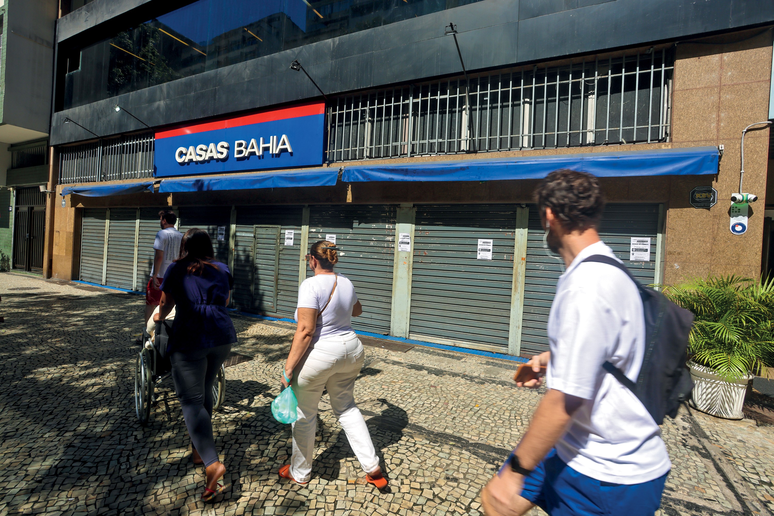 Pessoas caminham em calçada de paralelepípedos em frente a uma loja Casas Bahia com as portas de aço fechadas, indicando fechamento ou crise. Um homem de camiseta branca e mochila preta está à direita, uma mulher de blusa roxa empurra um carrinho e outra mulher de branco carrega uma sacola verde à esquerda.