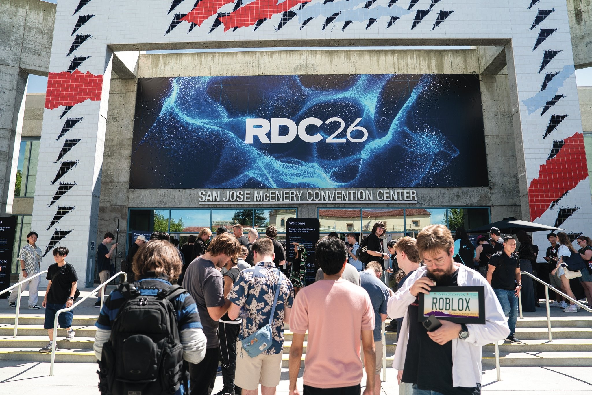 Pessoas em frente ao San Jose McEnery Convention Center, com um banner azul escuro exibindo RDC26 e um padrão de partículas luminosas. Um homem barbudo no primeiro plano segura um tablet com a palavra ROBLOX na tela. Outros participantes, alguns com mochilas, aguardam na entrada do evento