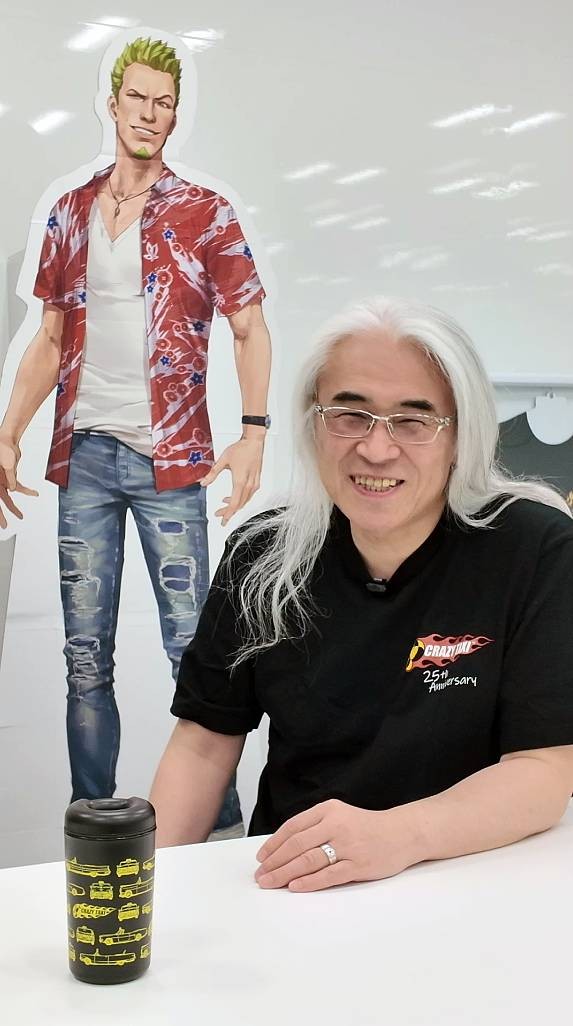 Homem asiático de cabelos longos e grisalhos, óculos, sorrindo, vestindo camiseta preta Crazy Taxi 25th Anniversary, sentado à mesa com um copo térmico preto estampado com carros amarelos. Atrás, um recorte de papelão de personagem masculino loiro, com barba, camisa havaiana vermelha e jeans rasgados.