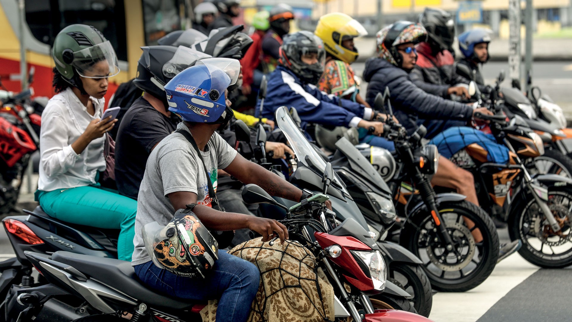 Grupo de motociclistas parados no trânsito. Uma mulher de capacete verde e blusa branca usa o celular, enquanto outros motociclistas, com capacetes variados, aguardam em suas motos.