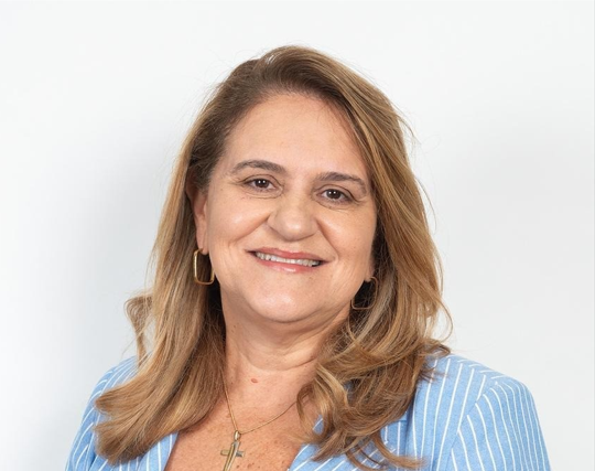 Mulher de meia-idade, cabelos loiros ondulados, sorri levemente para a câmera. Veste blazer azul claro listrado e colar com pingente de cruz dourada. Brincos argola retangulares dourados. Fundo branco.