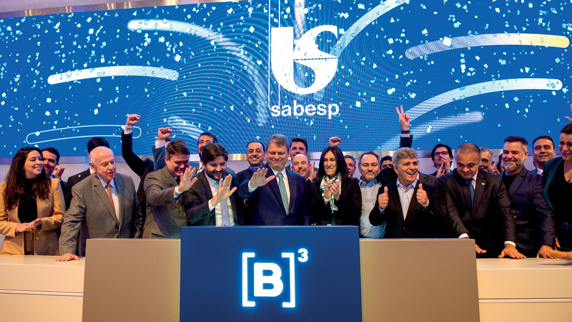 Grupo de pessoas sorrindo e acenando, algumas com os punhos erguidos, em frente a um telão azul com o logo da Sabesp e confetes digitais. À frente, um púlpito azul com o logo da B³.