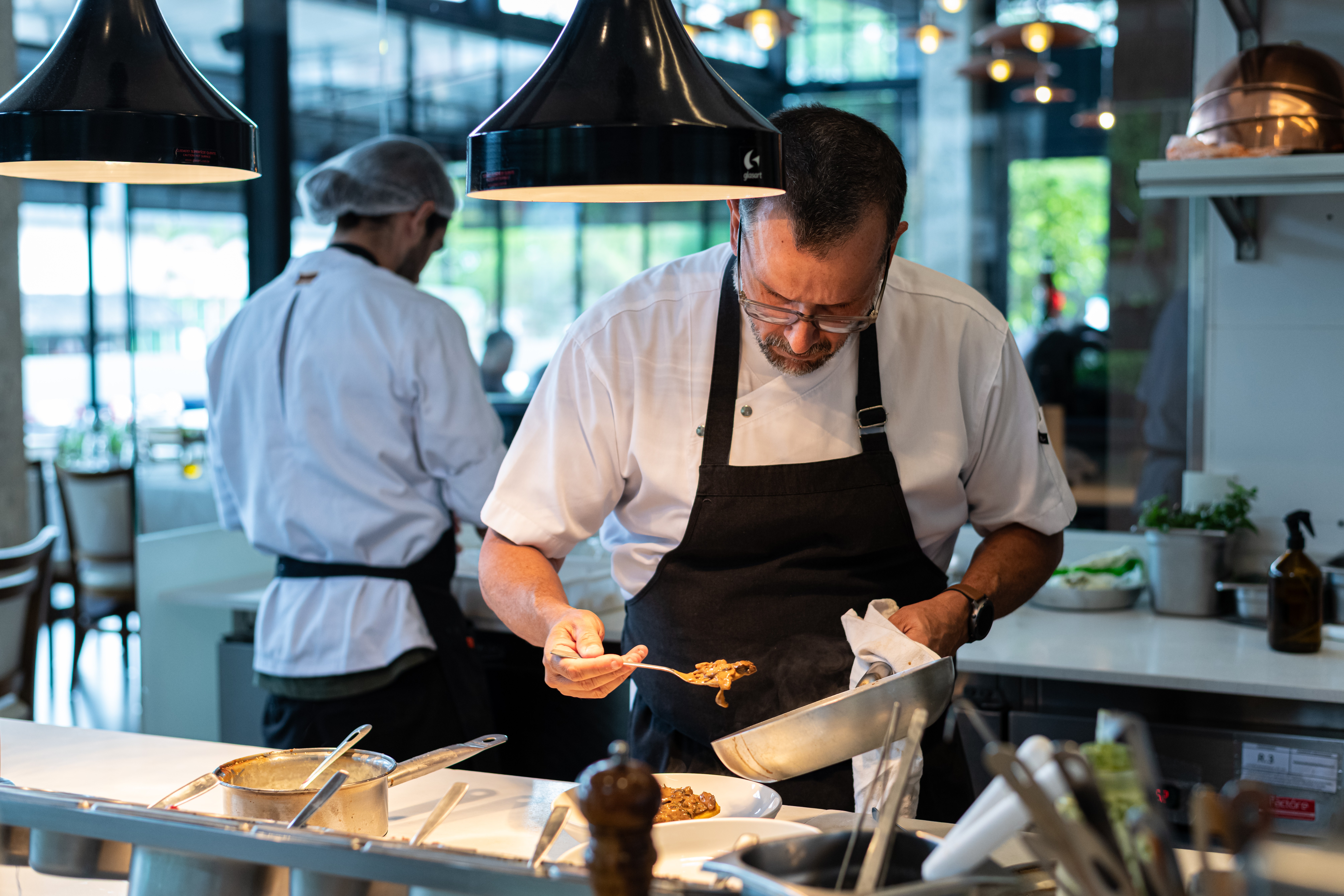 Chef de &oacute;culos e avental preto, concentrado, serve um prato com uma colher, segurando uma panela com um pano branco. Outro cozinheiro de touca e uniforme branco trabalha ao fundo, em uma cozinha moderna com ilumina&ccedil;&atilde;o pendente e detalhes em cobre