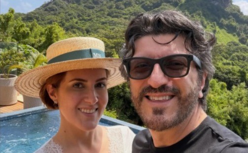 Casal sorridente, mulher de chapéu de palha e homem de óculos escuros, posando em frente a uma piscina com montanhas verdes ao fundo