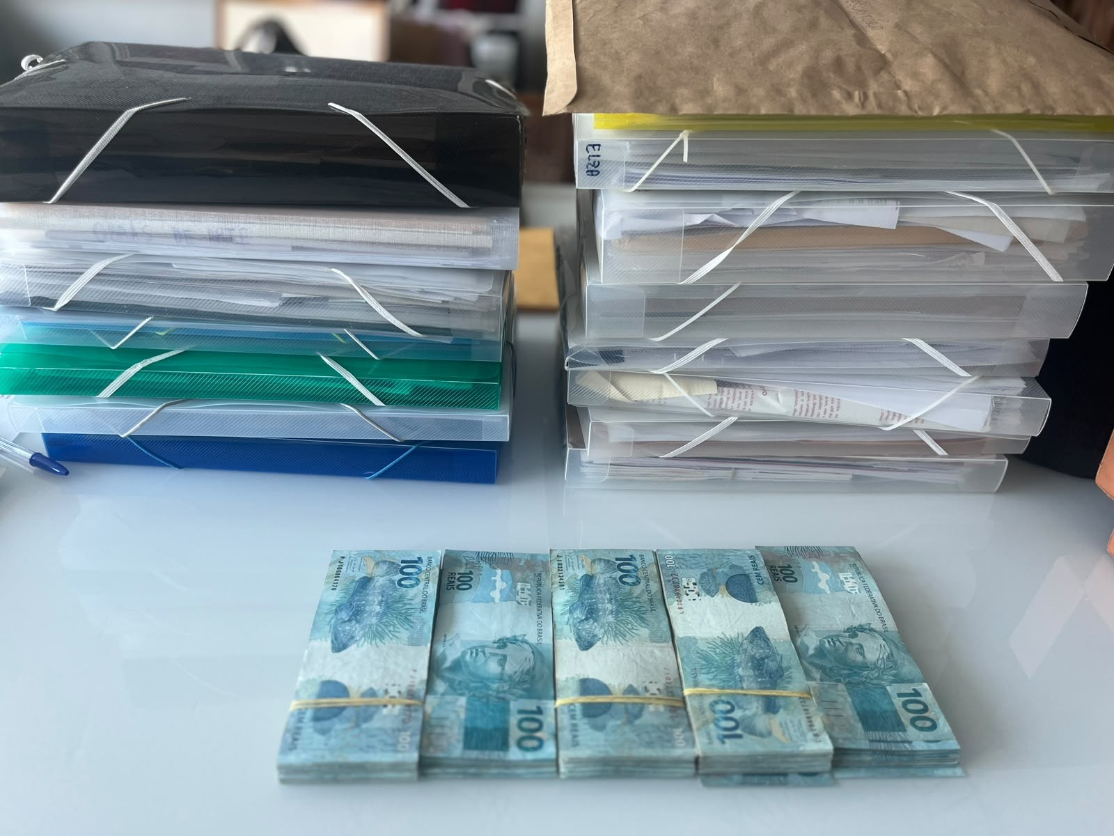 Cinco maços de notas de cem reais, presos por elásticos, sobre uma mesa branca, com pilhas de pastas de documentos ao fundo