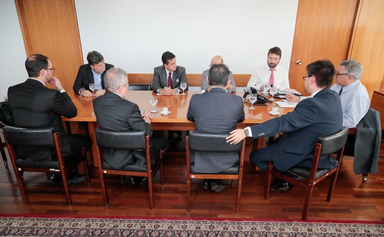 Oito homens de terno sentados em uma mesa de madeira retangular em uma reunião. Copos de água e xícaras de café estão sobre a mesa. O ambiente tem paredes claras e um tapete estampado no chão.