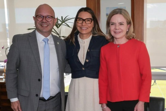 Três pessoas sorridentes posam para foto em ambiente interno. À esquerda, um homem careca de óculos, terno cinza e gravata azul clara. Ao centro, uma mulher de cabelos longos, óculos escuros, blusa branca e jaqueta jeans. À direita, uma mulher loira de casaco vermelho e calça preta. Ao fundo, uma planta e uma janela.