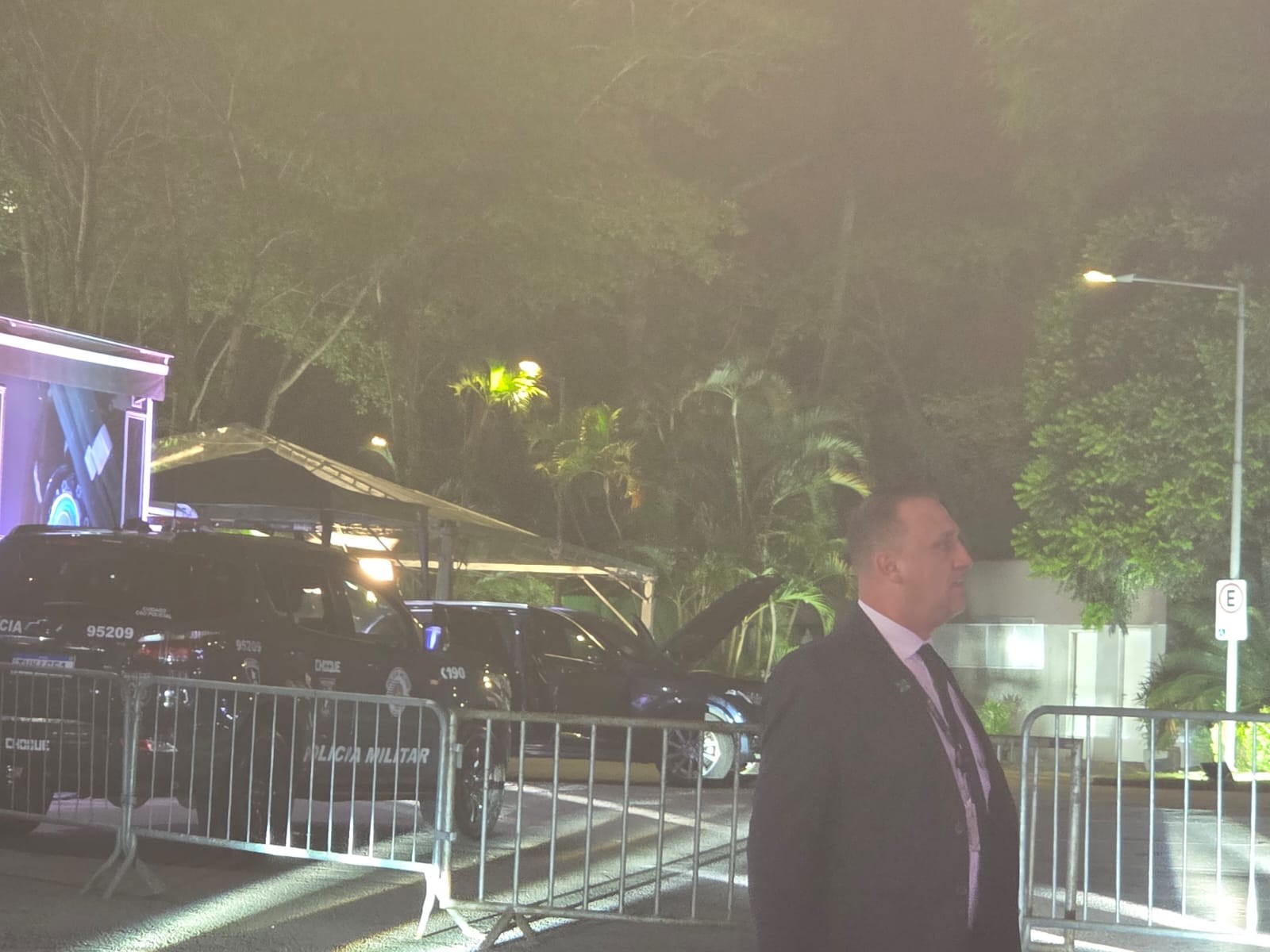 Homem de terno escuro e gravata, em perfil, olhando para a direita, em evento noturno com viaturas da Polícia Militar e grades de segurança. Ao fundo, árvores e iluminação artificial.