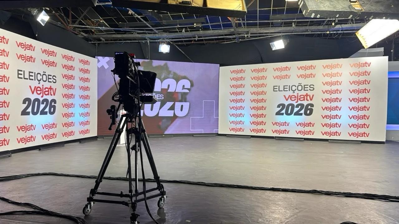 Estúdio de televisão com câmera profissional em tripé no centro, cabos pretos no chão. Ao fundo, painéis brancos exibem ELEIÇÕES Vejatv 2026 e o logo vejatv repetido. Um painel central roxo mostra texto ilegível