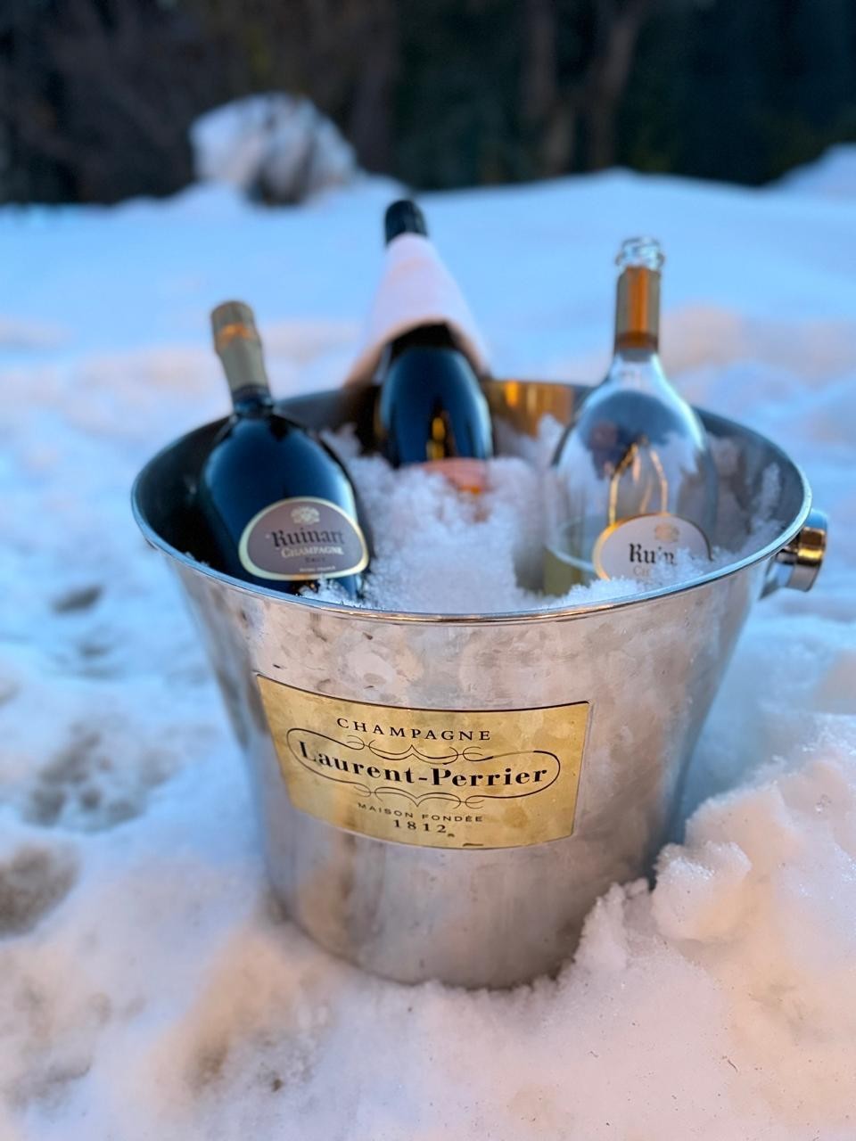 Balde de metal com três garrafas de champanhe e gelo, em meio a uma paisagem nevada. O balde tem um rótulo dourado com Champagne Laurent-Perrier Maison Fondée 1812.
