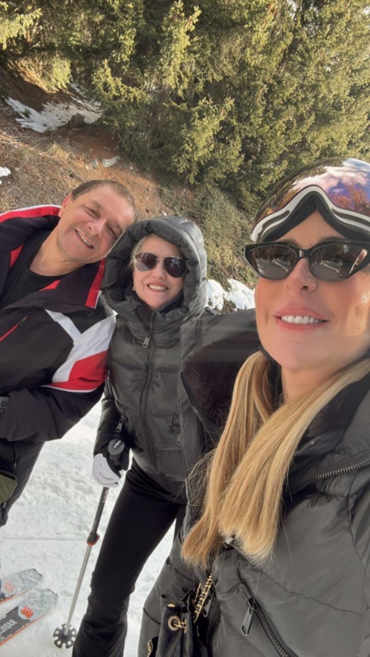 Três pessoas sorrindo em uma selfie na neve. Um homem à esquerda, uma mulher no centro com óculos de sol e capuz, e outra mulher à direita com óculos de sol e óculos de esqui na testa. Todos vestem roupas de inverno, com árvores e neve ao fundo