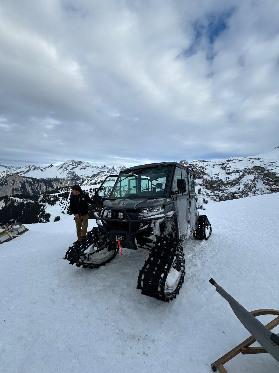 Veículo utilitário cinza com esteiras de neve estacionado em montanha coberta de neve. Um homem de jaqueta escura e calça clara está ao lado da porta aberta, olhando para a esquerda. Montanhas nevadas e céu nublado ao fundo.