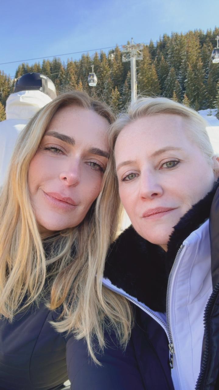 Duas mulheres loiras, uma com cabelos longos e outra com cabelos curtos, sorriem para a câmera em uma montanha nevada. Elas vestem casacos de inverno, e ao fundo há árvores cobertas de neve e teleféricos sob um céu azul