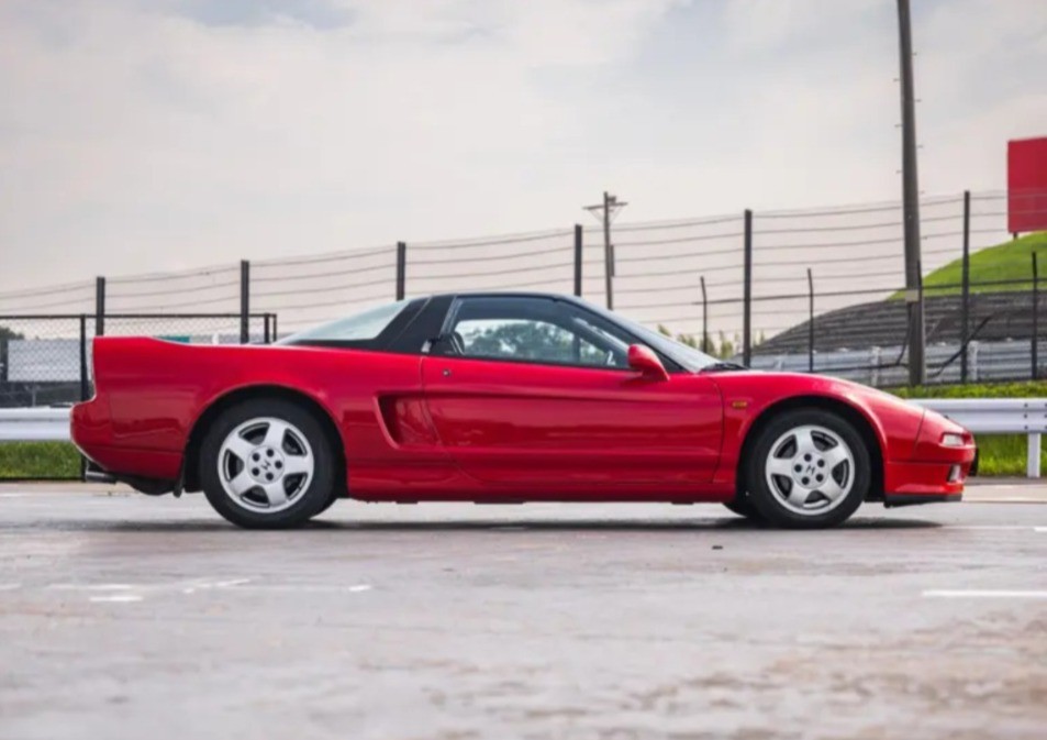 Carro esportivo vermelho, modelo Honda NSX, estacionado em uma pista de asfalto. O veículo tem teto preto e rodas prateadas. Ao fundo, uma cerca de proteção e um morro gramado sob céu nublado.