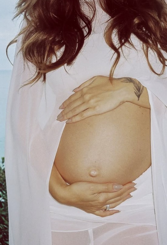 Mulher grávida com cabelos castanhos soltos, vestindo tecido branco transparente, segura a barriga com as duas mãos. Uma tatuagem de pena está visível no dorso da mão esquerda e um anel prateado no dedo anelar direito. O umbigo está saliente