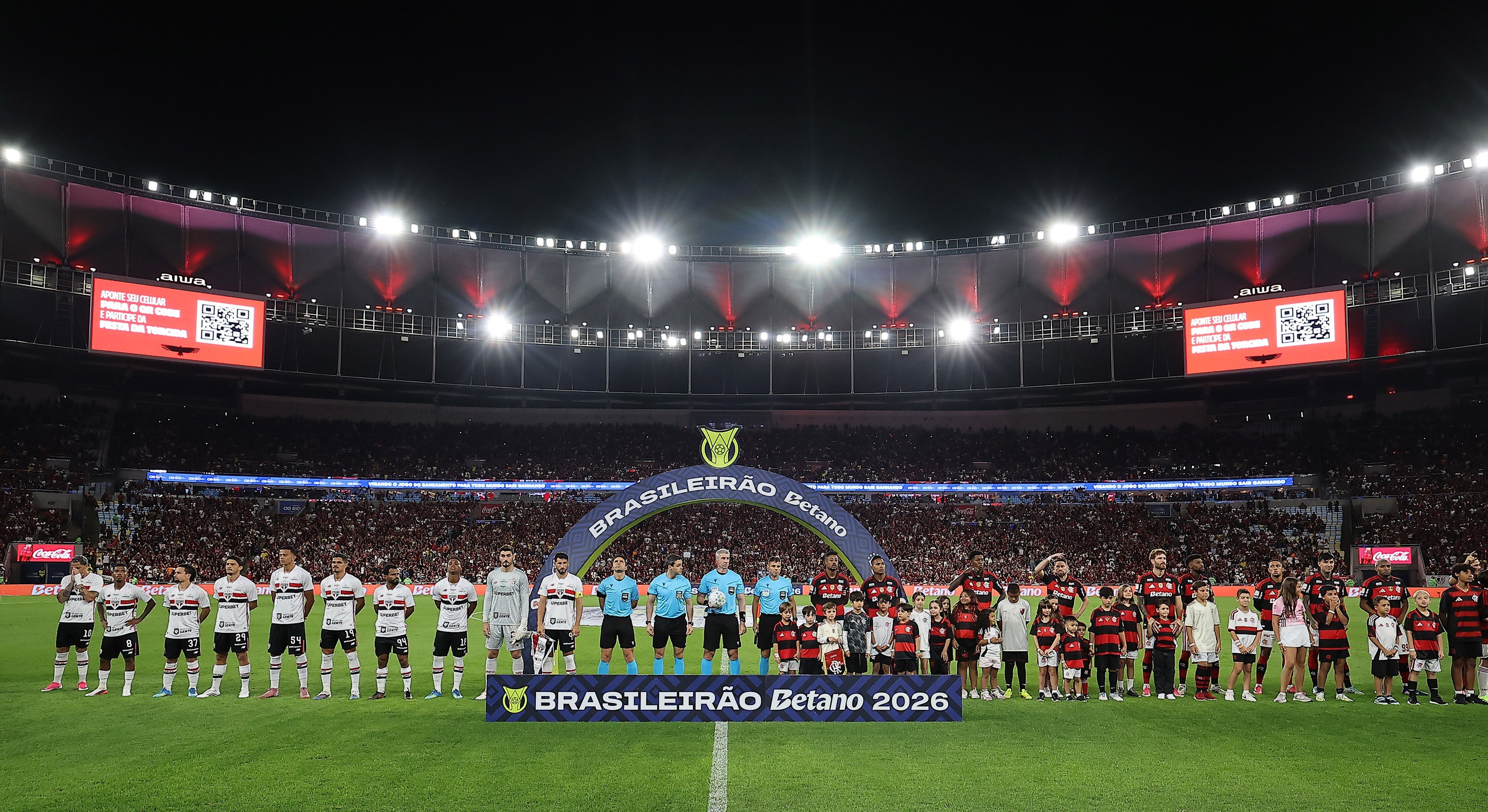 Jogadores de futebol e crianças perfilados no campo de um estádio lotado à noite, com telões exibindo QR codes e a inscrição Brasileirão Betano 2026 em um banner