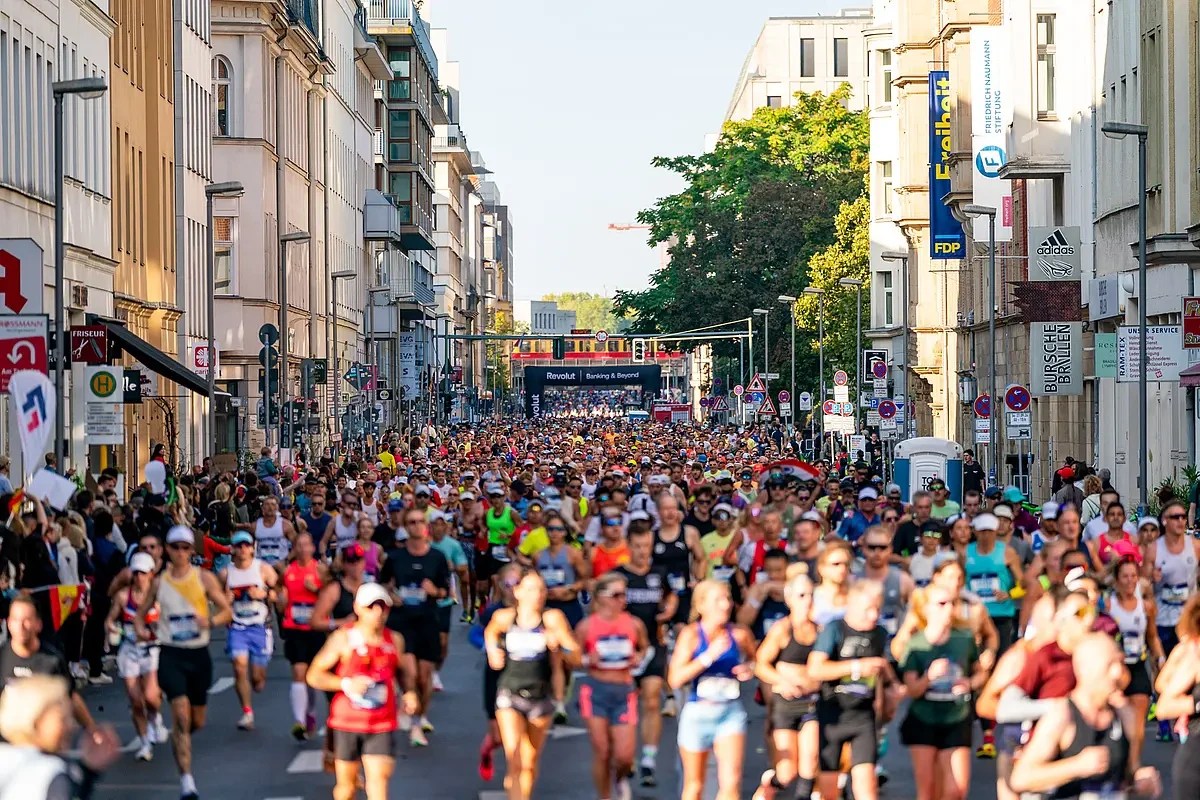 Multidão de corredores de maratona, homens e mulheres de diversas idades, correndo em uma rua larga de cidade, ladeada por prédios comerciais e residenciais. Ao fundo, um pórtico preto com a inscrição Revolut Running & Beyond marca o início ou fim da corrida. O céu está claro, e a luz do sol ilumina a cena, criando um clima vibrante e energético.