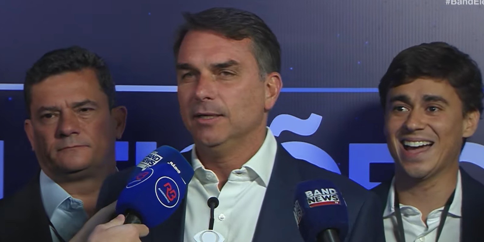 Jair Bolsonaro, de terno azul e camisa branca, fala em microfones da Band News e Rádio Bandeirantes, com Sérgio Moro à esquerda e um jovem sorridente à direita, em evento político