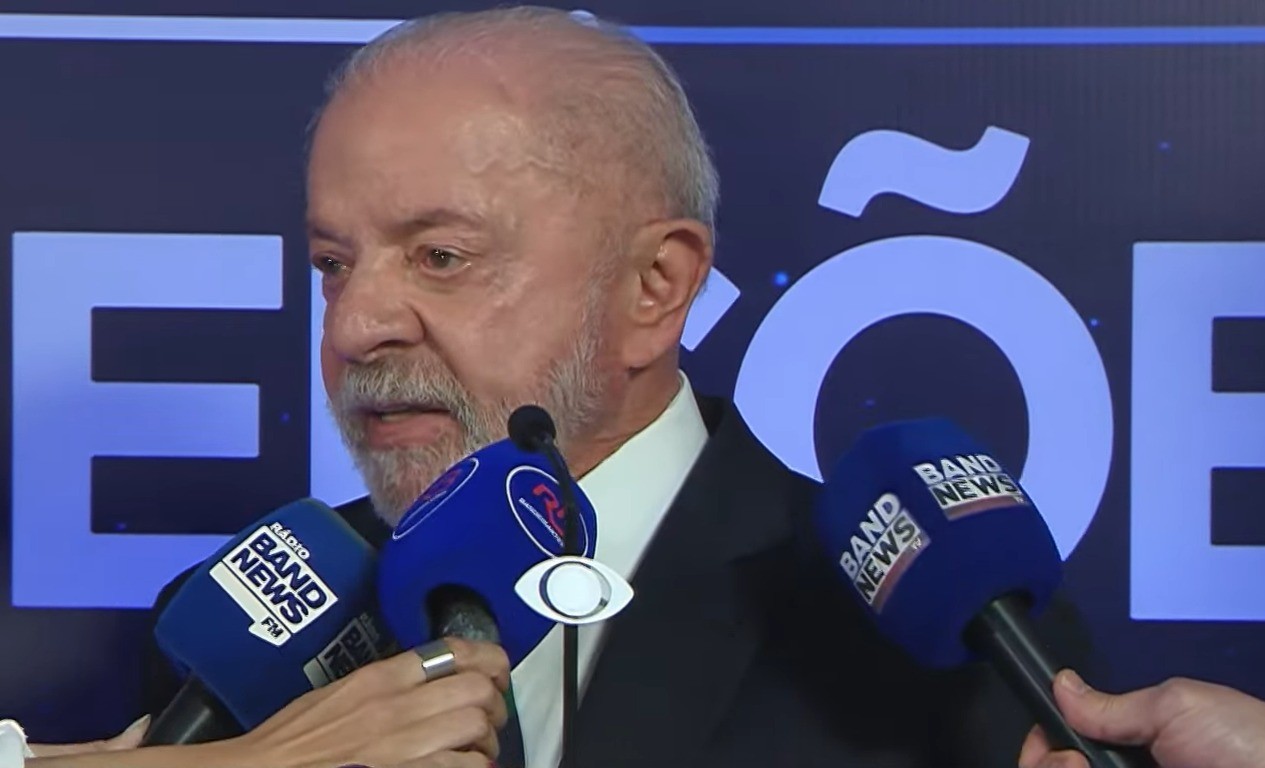 Homem idoso de cabelos e barba brancos, vestindo terno escuro e camisa branca, fala em microfones azuis da Band News. Ele está em um ambiente escuro com letras grandes e claras ao fundo