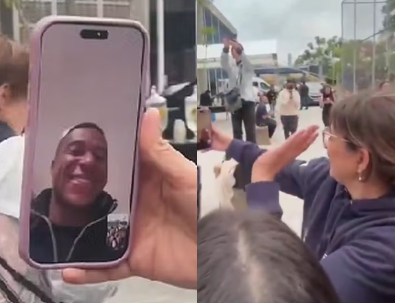 Mão segura um celular com um homem negro sorridente em videochamada, enquanto uma mulher de óculos e blusa azul acena para a tela, em meio a uma multidão em ambiente externo