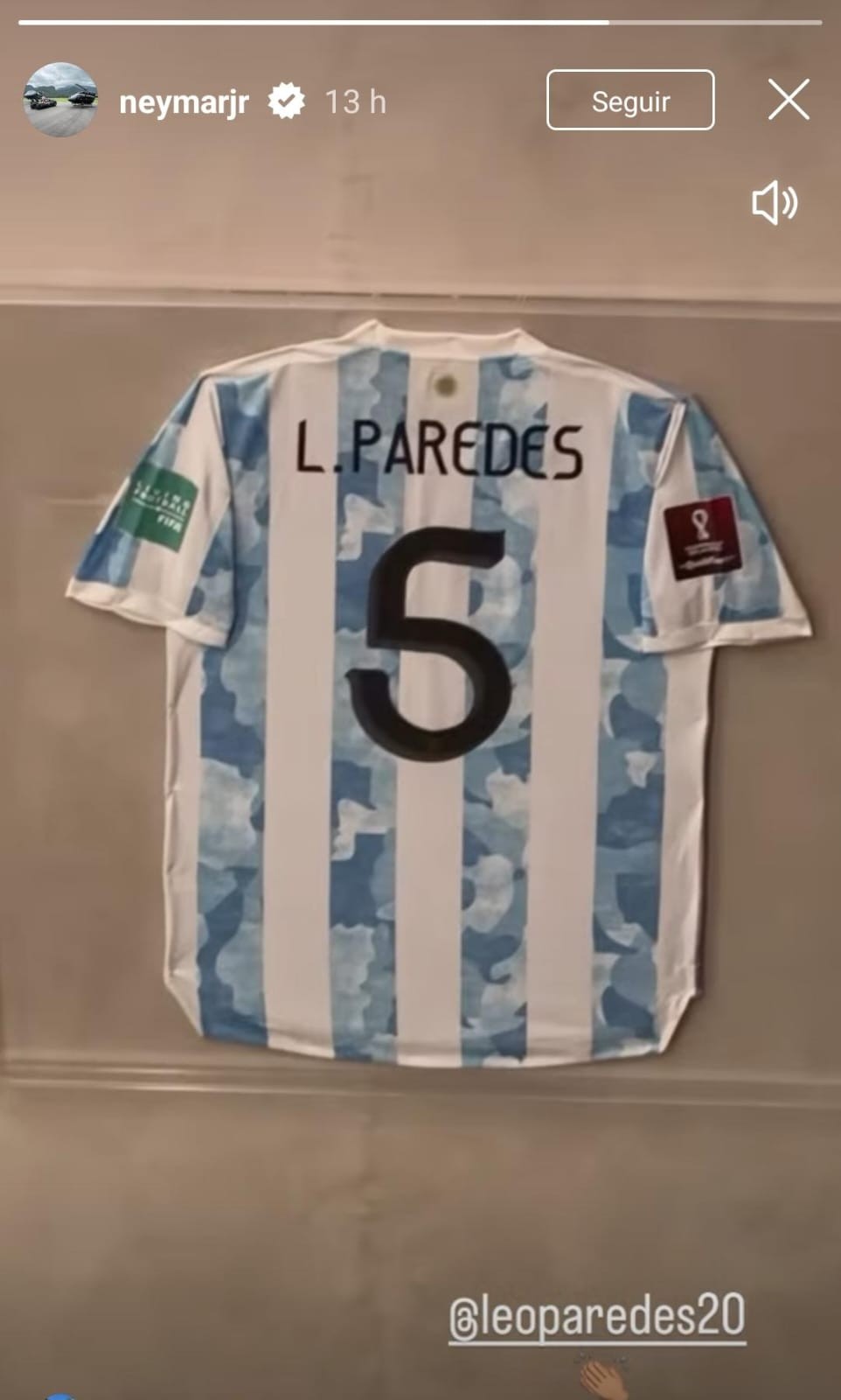 Camisa da seleção argentina de futebol, branca e azul-claro listrada, com o nome L. PAREDES e o número 5 em preto nas costas. Há um emblema da FIFA na manga esquerda e um da Copa do Mundo na direita. A camisa está pendurada em uma parede cinza-clara. Na parte superior da imagem, há elementos de interface de usuário de uma rede social, incluindo o nome de usuário neymarjr e um ícone de verificação, e na parte inferior, a menção @leoparedes20