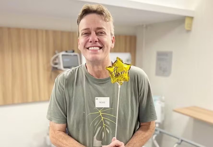 Homem loiro de meia-idade, sorrindo, usando camiseta verde-oliva com estampa de folha, segurando um balão dourado em formato de estrela com a palavra Sim escrita. Ele está em um ambiente claro, com paredes brancas e detalhes amadeirados, parecendo um hospital ou clínica