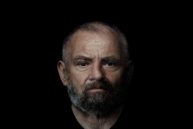 Rosto de um homem de meia-idade, calvo no topo da cabeça, com barba e bigode grisalhos. Ele tem uma expressão séria e olha diretamente para frente, com a pele marcada por rugas. A iluminação destaca seu rosto contra um fundo escuro