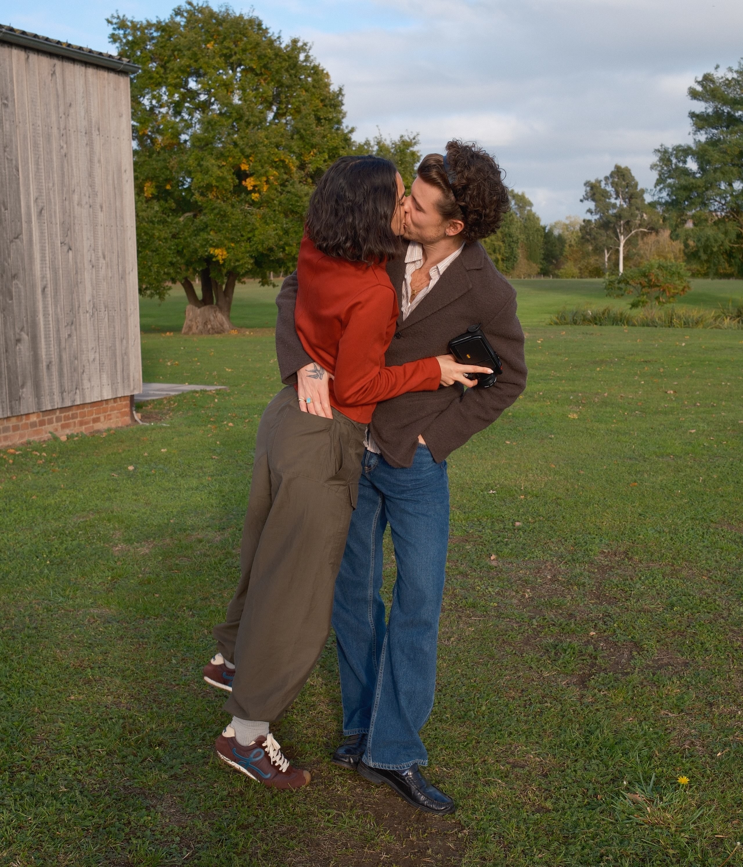 Casal jovem se beijando apaixonadamente em um gramado verde. A mulher, de cabelo escuro e blusa terracota, abraça o homem, que veste jaqueta marrom e jeans. Ele segura uma carteira preta. Ao fundo, árvores e um céu azul com nuvens.